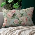 A rectangular floral throw pillow with pink roses on a pale blue-green background, styled on a light gray sofa with a cream knit blanket and a dark green cushion behind it.