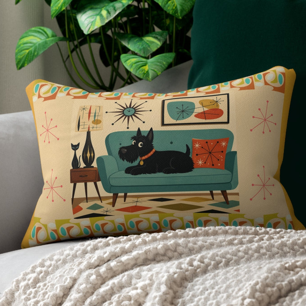 A rectangular decorative throw pillow with a mid-century modern illustration of a black cat on a teal sofa, styled on a light gray sofa with a cream knit blanket and a green potted plant in the background.